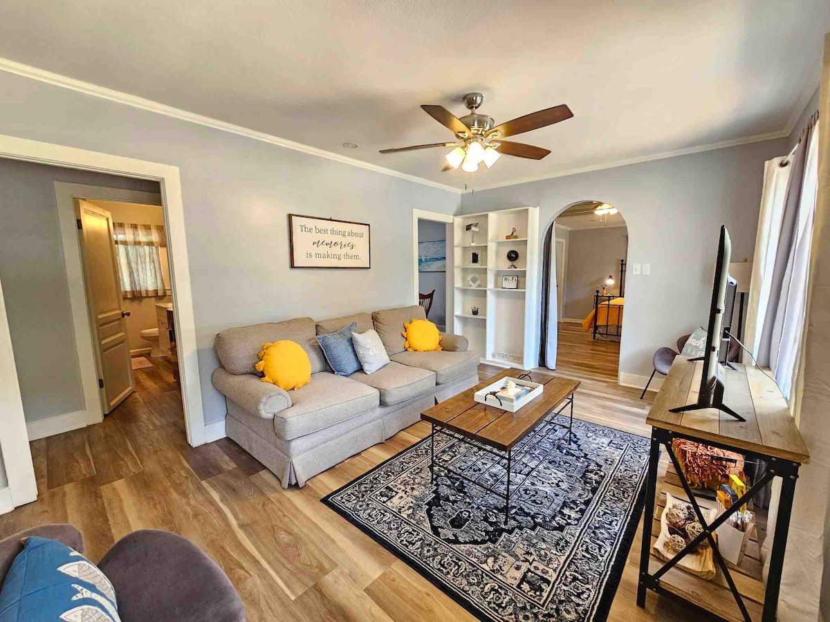 A cozy living room is showcased, featuring a snug sofa adorned with colorful pillows and a stylish coffee table positioned on a patterned area rug. A ceiling fan adds a touch of comfort, while sunlight pours in through the windows, illuminating the welcoming space.