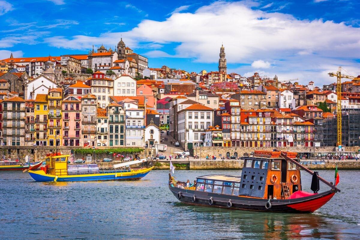The image showcases a vibrant riverside scene in Porto, with colorful buildings stacked on a hillside under a partly cloudy sky. Two boats are floating on the river, one featuring a traditional design with dark wood and decorative elements, the other painted in bright yellow and blue.