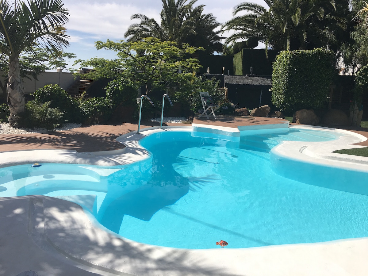 A serene swimming pool features crystal-clear water, surrounded by lush greenery and palm trees. A sun lounger is positioned nearby, and natural stone accents complement the landscaping, creating an inviting outdoor space for relaxation.