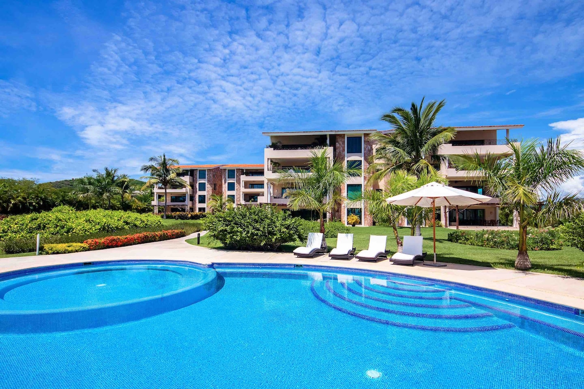 The image showcases a vibrant pool area, featuring a circular swimming pool with clear blue water. Four sun loungers are positioned nearby under a white umbrella, surrounded by lush greenery and tropical plants. The backdrop includes a multi-story building, reflecting the inviting atmosphere of the community.