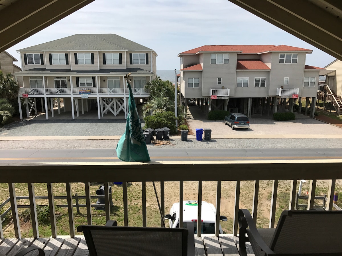 A view is captured from a balcony, overlooking two neighboring beach houses. The houses are elevated, with palm trees and grassy areas visible nearby. A car is parked on the road, with trash bins positioned alongside, providing a glimpse of the quiet street.
