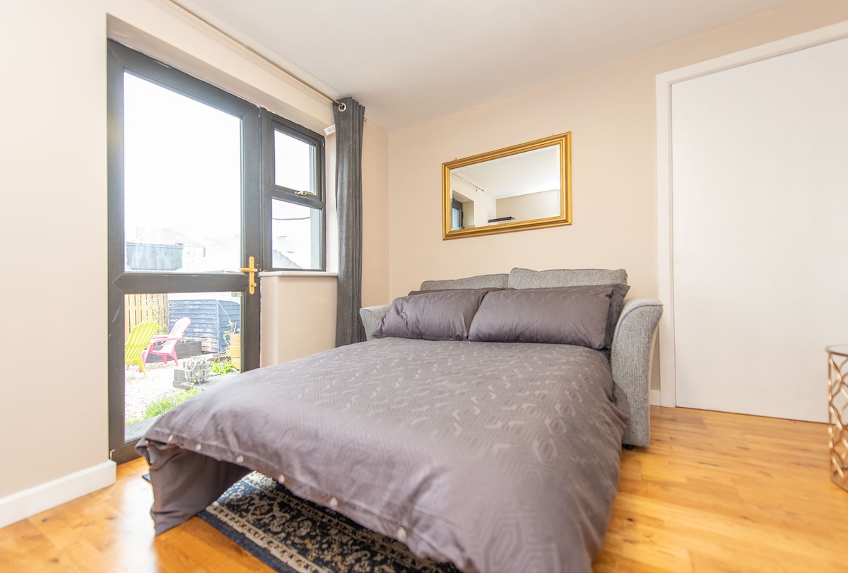 A spacious sleeping area is furnished with a cozy grey couch positioned beside large glass doors that offer access to a private garden. A decorative mirror hangs on the wall, reflecting natural light, while warm wooden flooring complements the overall design.