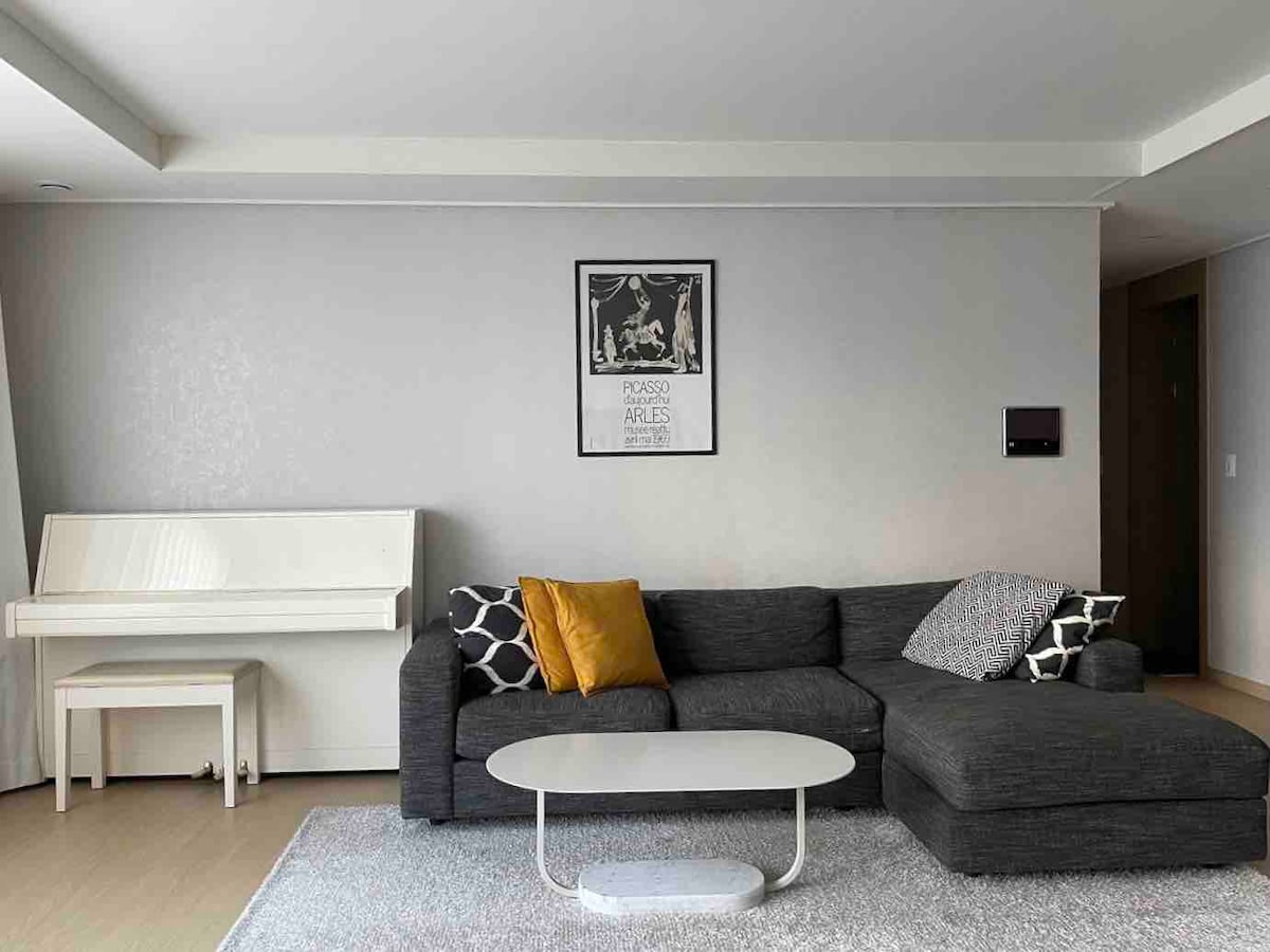 A spacious living area is characterized by a dark sectional sofa adorned with patterned cushions. A sleek coffee table is positioned in front of the sofa on a light-colored rug. A white piano stands against a neutral wall, complemented by decorative wall art.