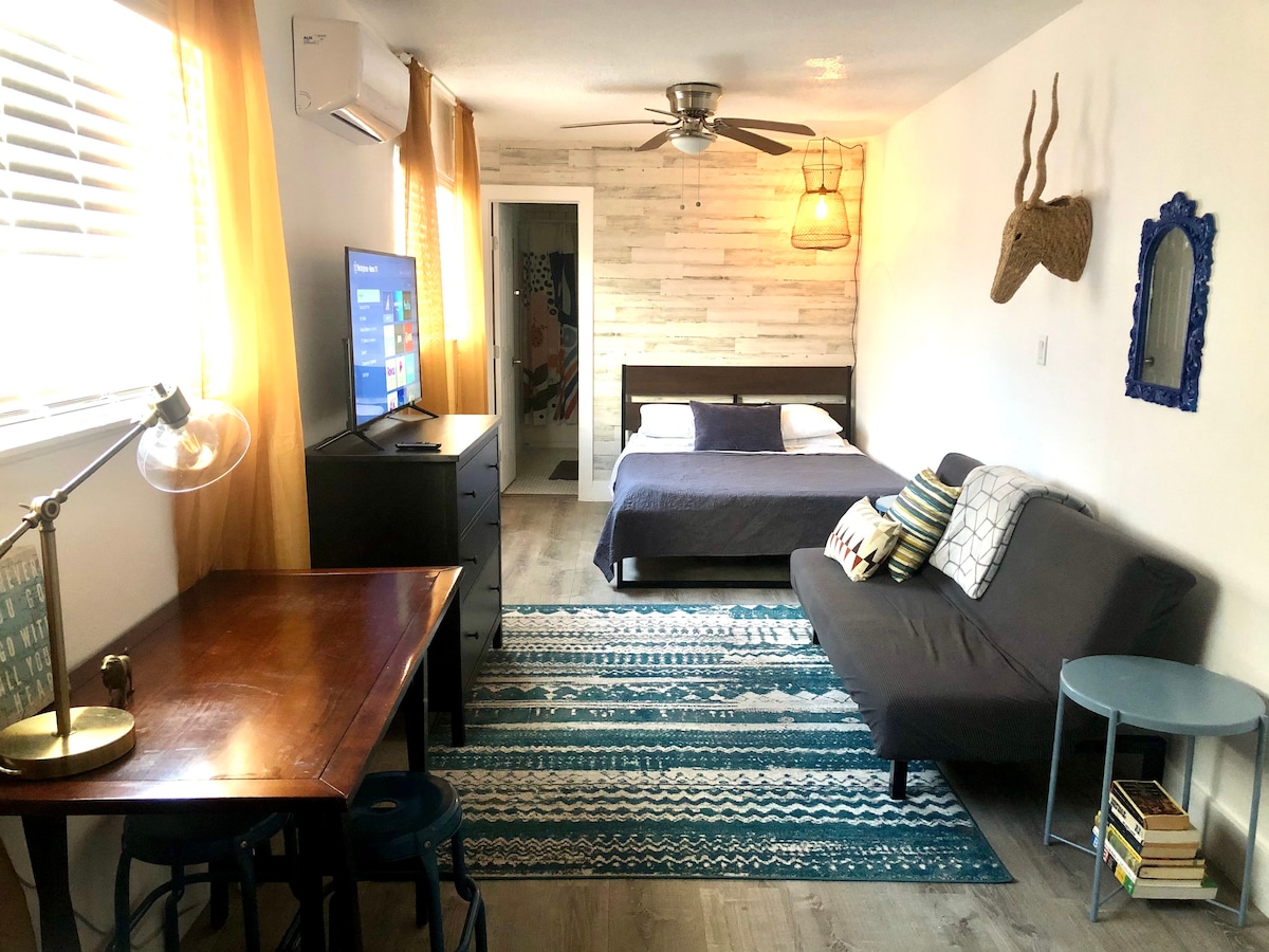 The studio apartment features a queen-size bed and a short twin futon, positioned on a wooden floor. A dining table with chairs is nearby. A television and a decorative mirror are present, with warm light filtering through sheer curtains. A cozy rug adds texture to the space.