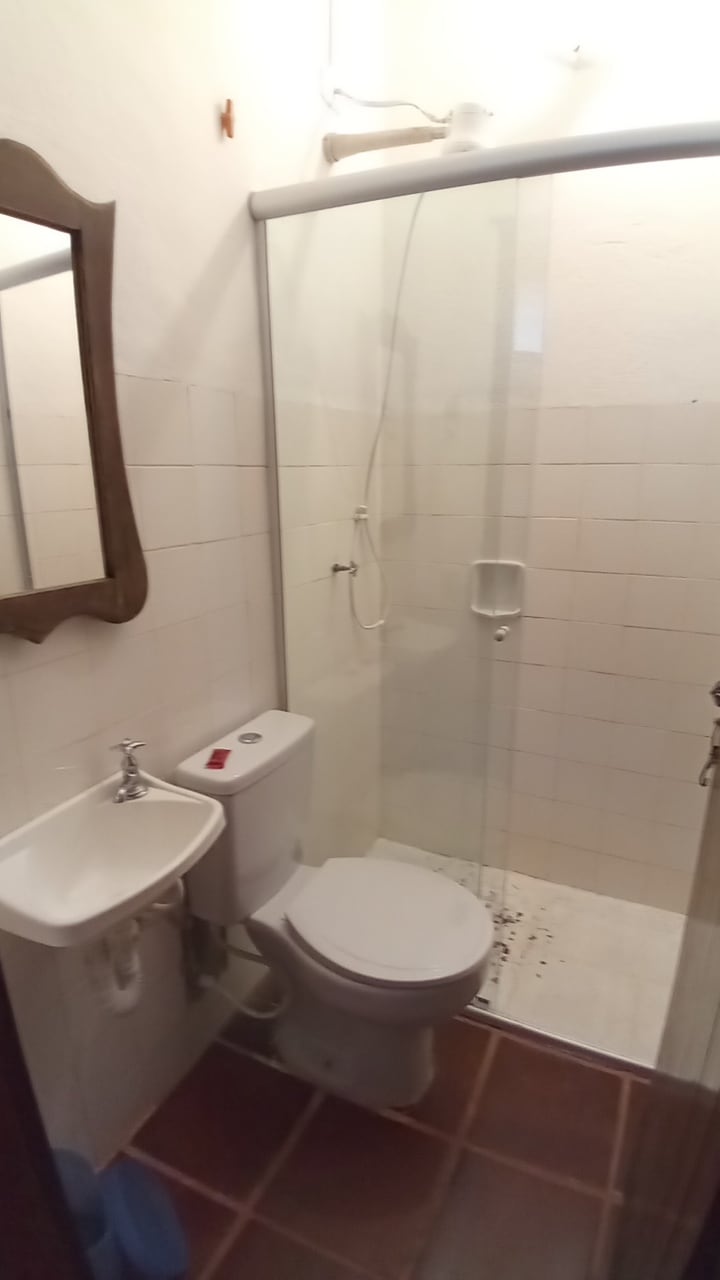 A compact bathroom features a clear glass shower enclosure, a white toilet, and a small sink. A mirror with a wooden frame is mounted on the wall. The tiled floor adds a clean and functional touch to the space.