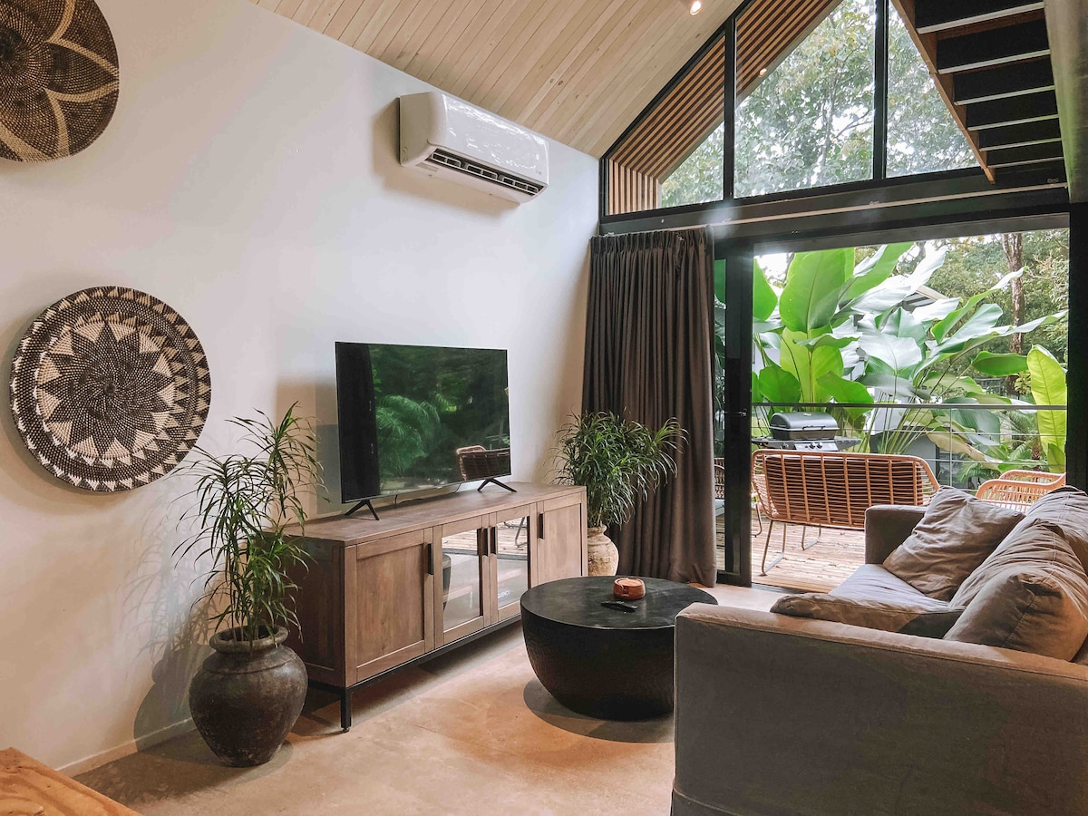 The living area features a comfortable sofa and a modern television on a wooden console. Large windows allow natural light to fill the space, while views of tropical greenery are visible outside. Decorative elements include plants and woven wall art.