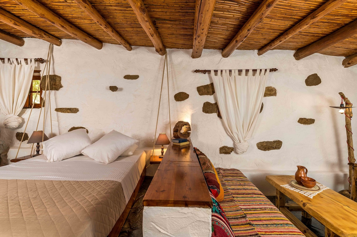 A spacious sleeping area features a large double bed with crisp linens, complemented by decorative pillows. Natural light filters through a window adorned with light curtains. A rustic table runs along the wall, accompanied by two wooden benches and colorful textiles, enhancing the inviting ambiance.