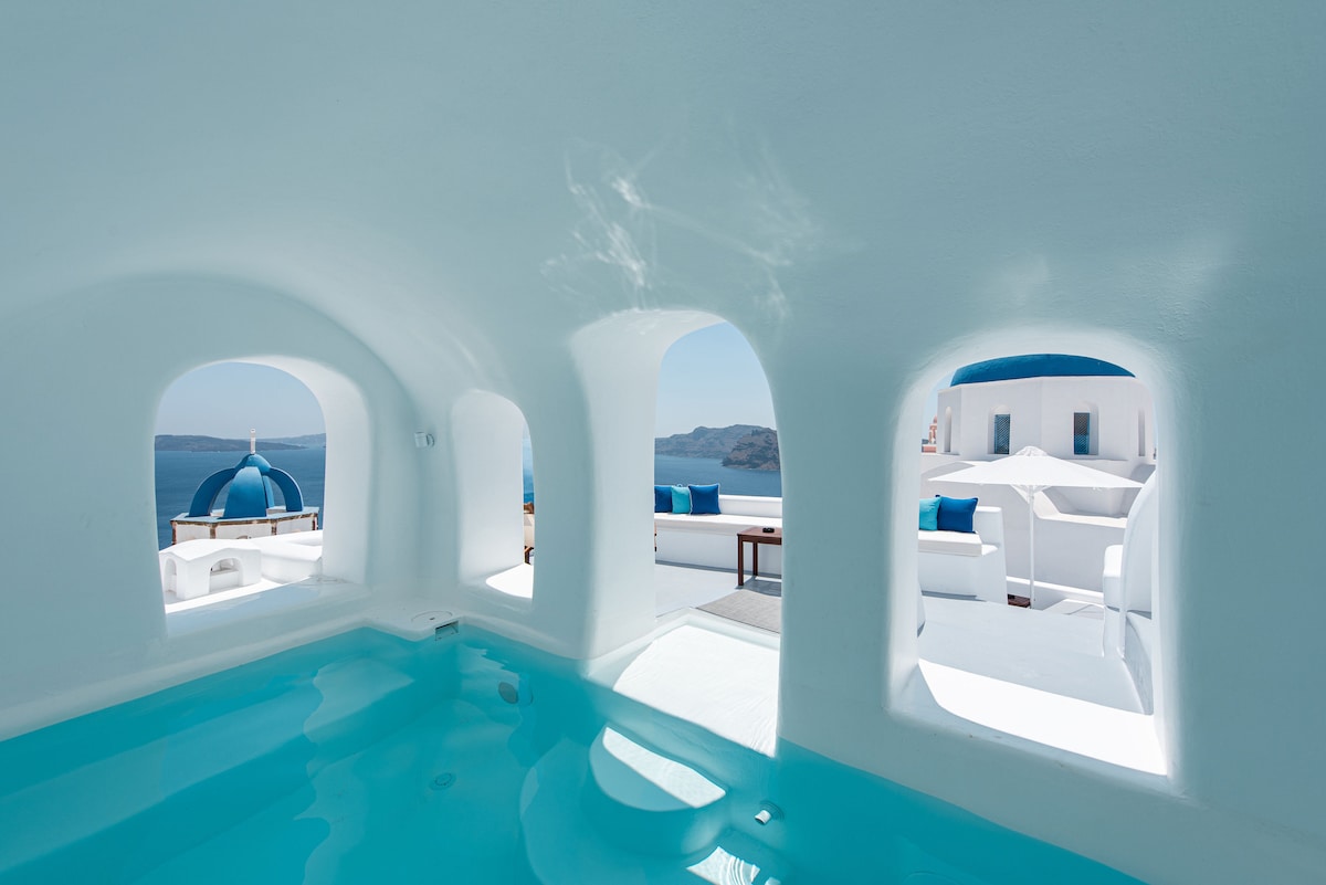 A serene cave pool area is framed by three arched windows, allowing natural light to illuminate the space. The inviting turquoise water reflects soft tones, while a seating area outside features blue cushions and a white umbrella, complemented by stunning views of the caldera.