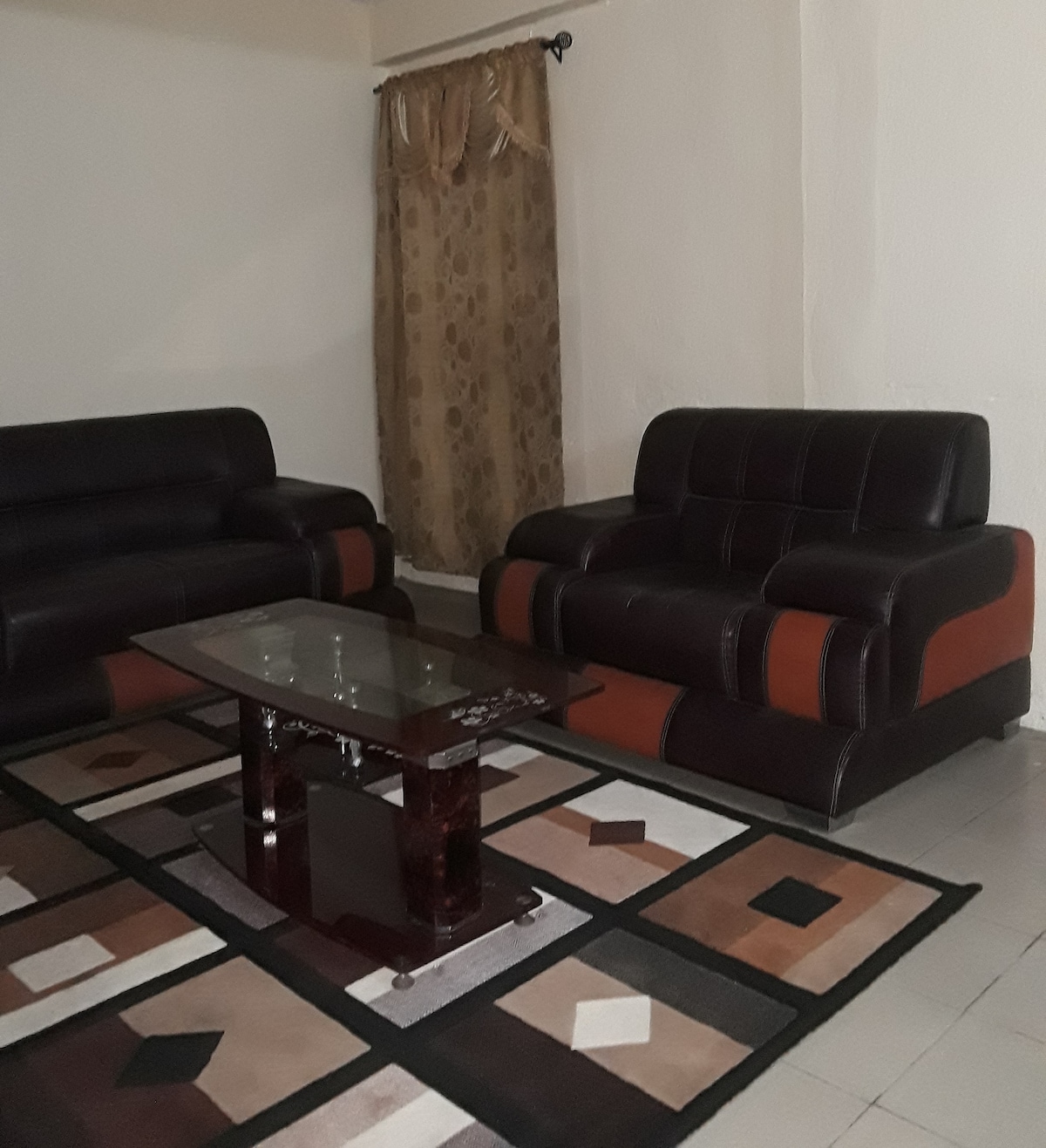 A comfortable seating area features a pair of dark leather sofas arranged around a rectangular glass-topped table. A textured rug with geometric patterns lies beneath, while a curtain with a subtle design frames a nearby window.