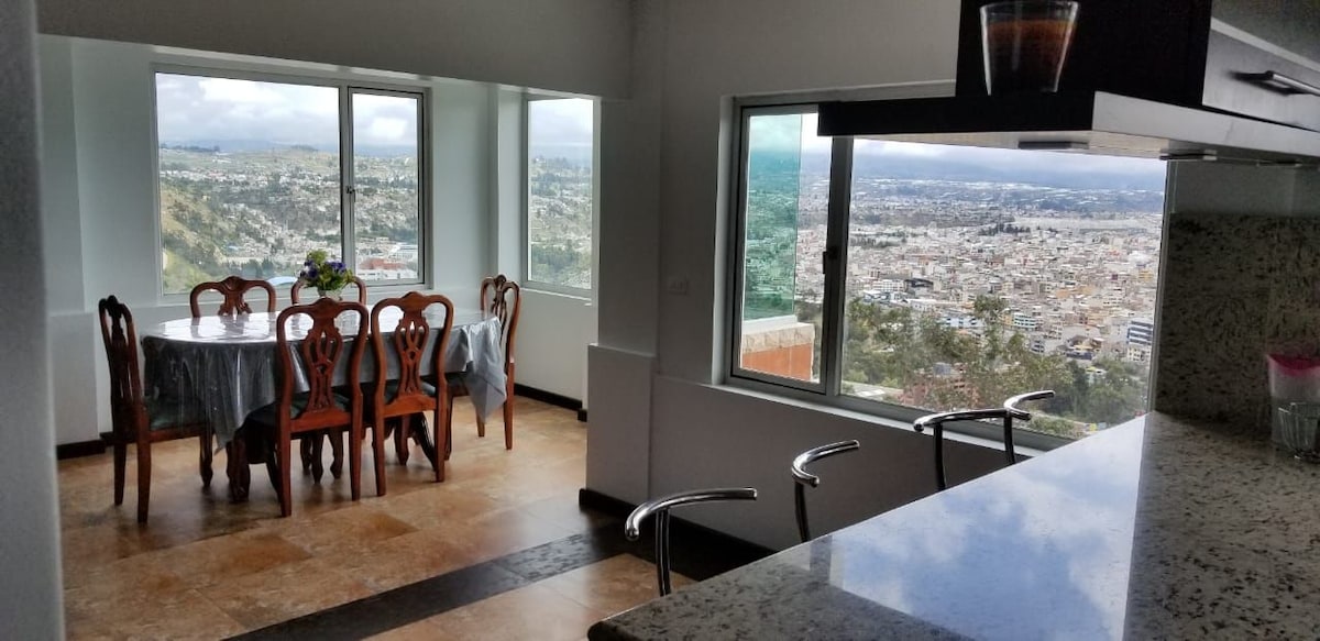 The dining area features a wooden table surrounded by chairs, positioned near large windows offering a panoramic view of Ambato. Counter space with bar stools is visible in the foreground, highlighting the open layout of the space.