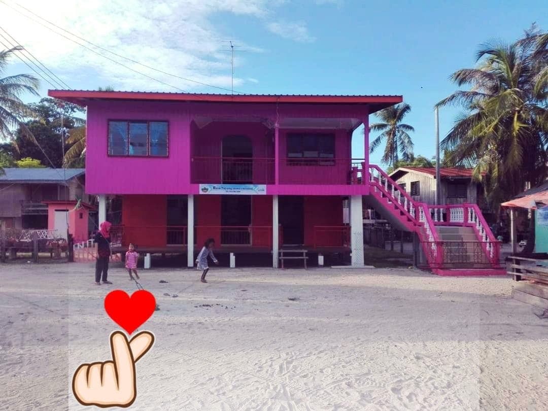A vibrant two-story structure is presented in bright pink, set against a clear blue sky. The building features an inviting staircase leading to a balcony, surrounded by palm trees. A sandy area in front allows for outdoor play, with children visible engaging in activities.