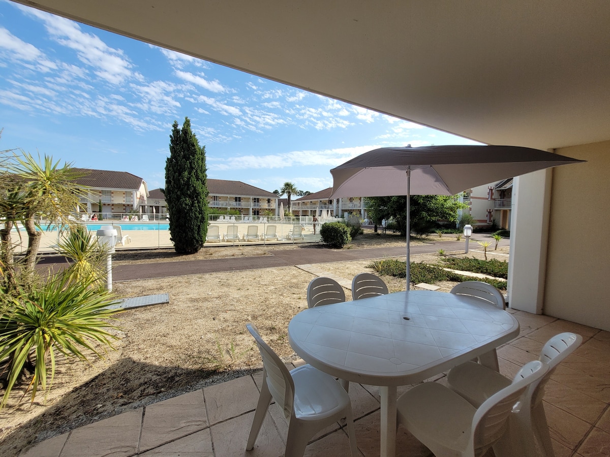 An outdoor terrace is depicted with a round table and six white chairs, shaded by a large umbrella. In the background, a clear view of the pool area is visible, surrounded by landscaped gardens and buildings, under a partly cloudy sky.