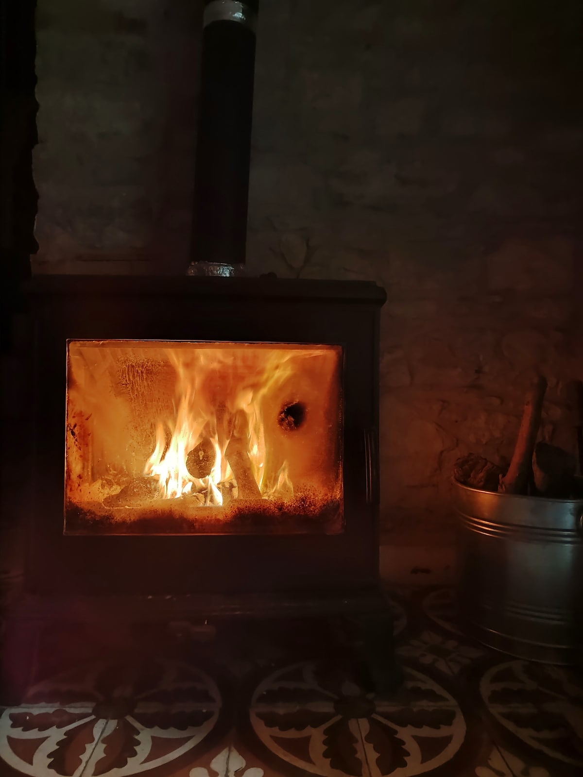 A wood-burning stove is seen emanating warm flames, set against a rustic stone wall. The flickering firelight casts a soft glow, highlighting the cozy ambiance. A metallic bucket with logs is positioned nearby, enhancing the inviting scene.