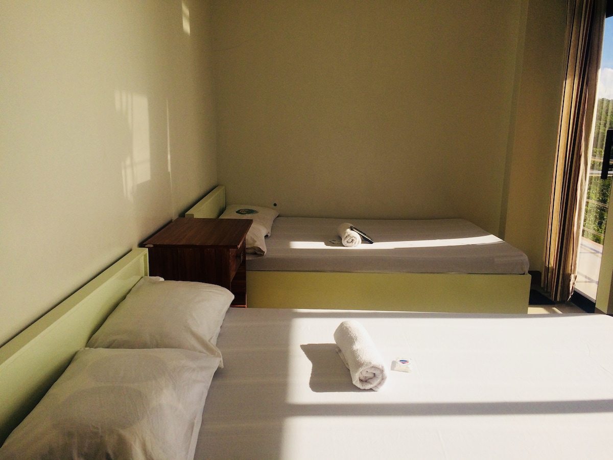 A bright sleeping area features two beds, one made with white linens and an additional unmade bed beside it. A wooden nightstand is positioned next to the made bed, and sunlight filters in through a nearby window, creating a warm ambiance.