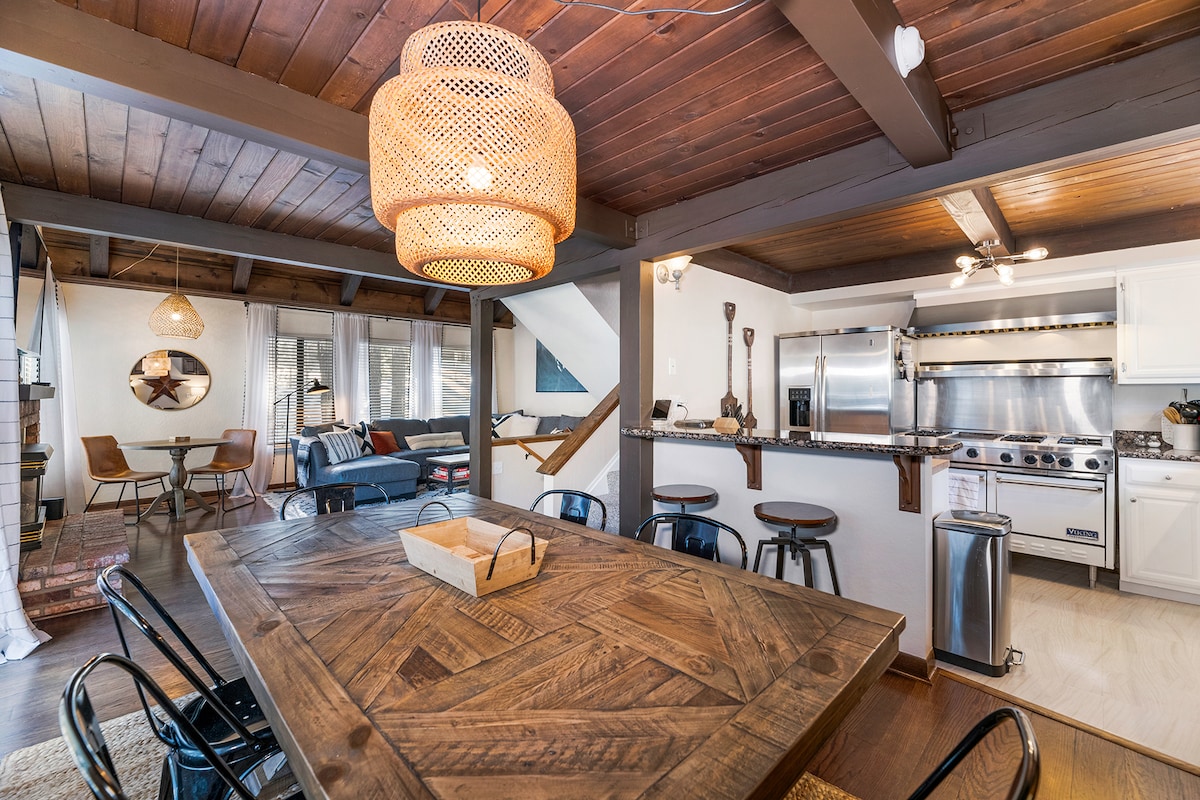 The dining area features a wooden table surrounded by black chairs, illuminated by a woven pendant light. A cozy living space with a sectional sofa is visible in the background, along with a well-equipped kitchen showcasing stainless steel appliances and a breakfast bar.