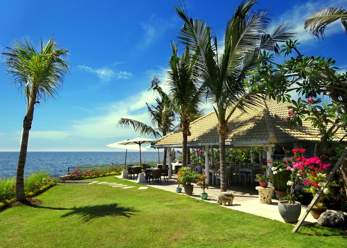 A spacious lawn area is bordered by vibrant tropical plants and palm trees, leading to a beachfront terrace. A charming building features a thatched roof, with outdoor seating under shaded umbrellas, offering unobstructed views of the ocean and clear blue sky.