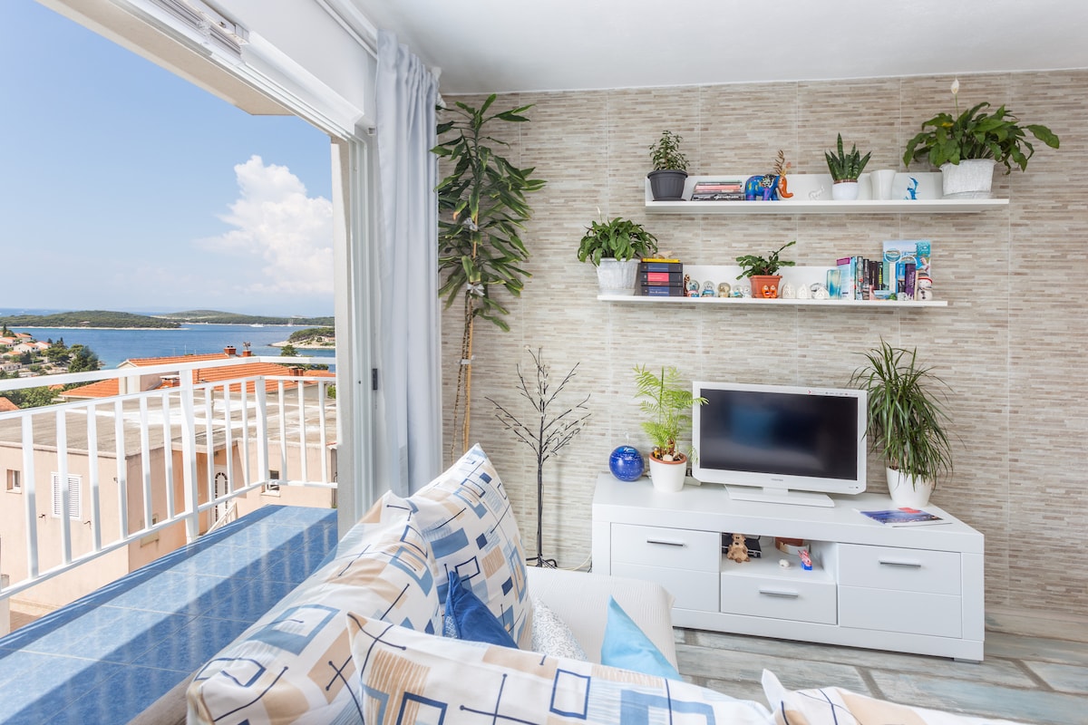 A bright living room is presented, featuring a white TV stand with decorative plants and books on the shelves. A comfortable sofa with patterned cushions offers views through large glass doors that lead to a balcony overlooking the sea and nearby landscape.