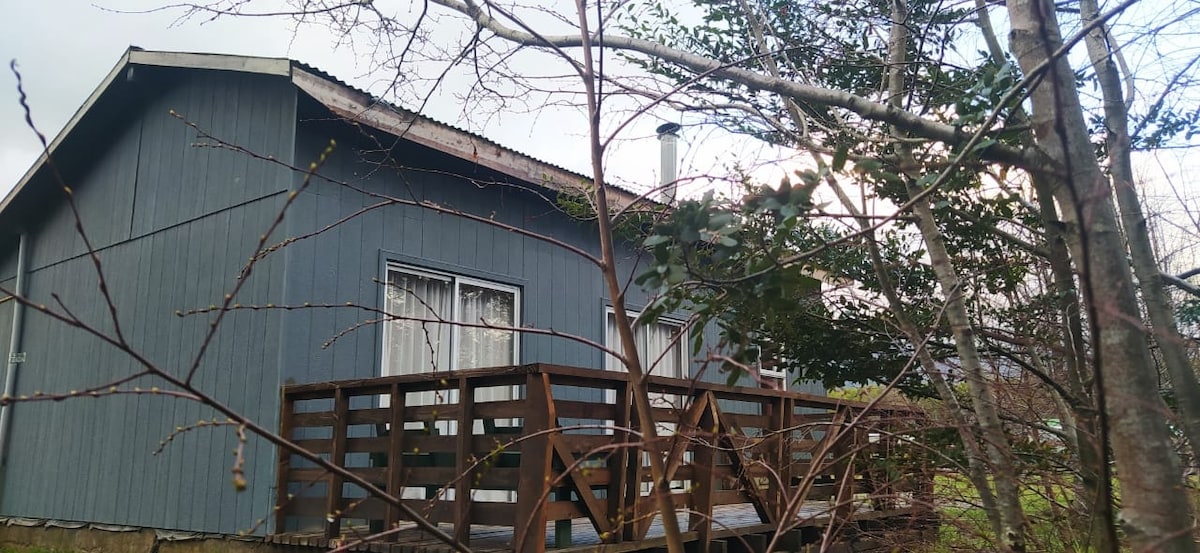 A wooden cabin is nestled among trees, showcasing a deck made of natural wood. Large windows are visible, allowing light to enter the interior. The building is surrounded by greenery, highlighting its connection to nature.