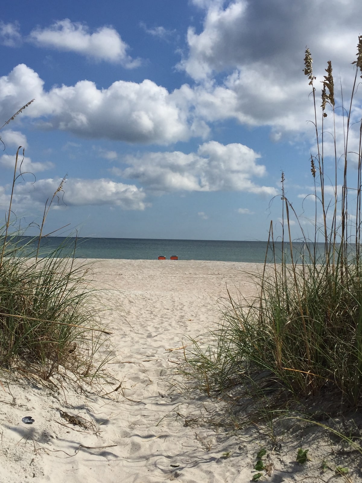 A sandy pathway leads to a quiet beach, framed by tall grasses on either side. The calm ocean extends toward the horizon under a partly cloudy sky. Two bright orange chairs are visible in the distance, inviting relaxation by the water.
