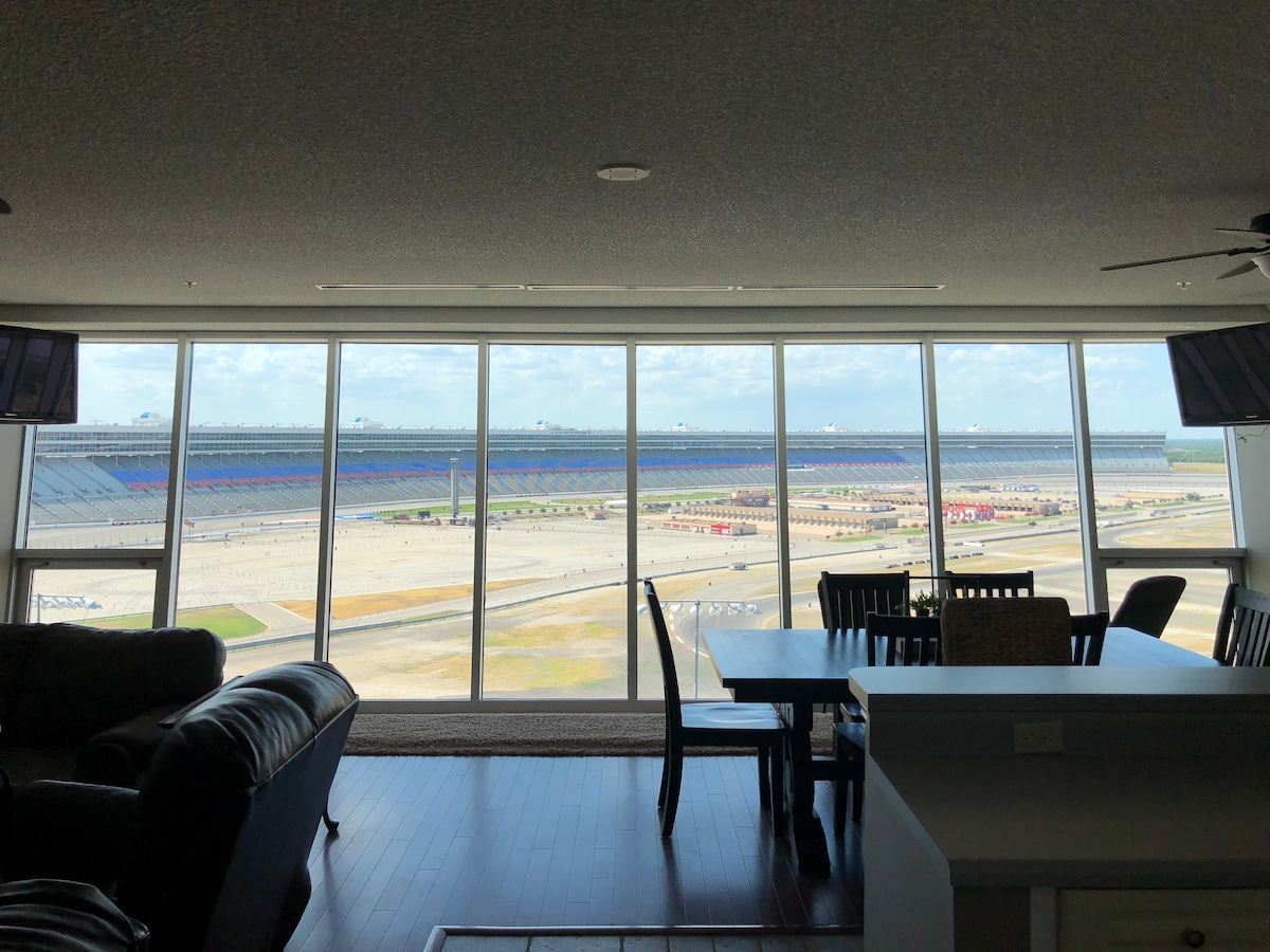 A spacious living area features a large wall of windows offering unobstructed views of the speedway. The seating area includes a black leather couch and dining table, with natural light illuminating the space. The expansive view showcases the racetrack and surrounding area.