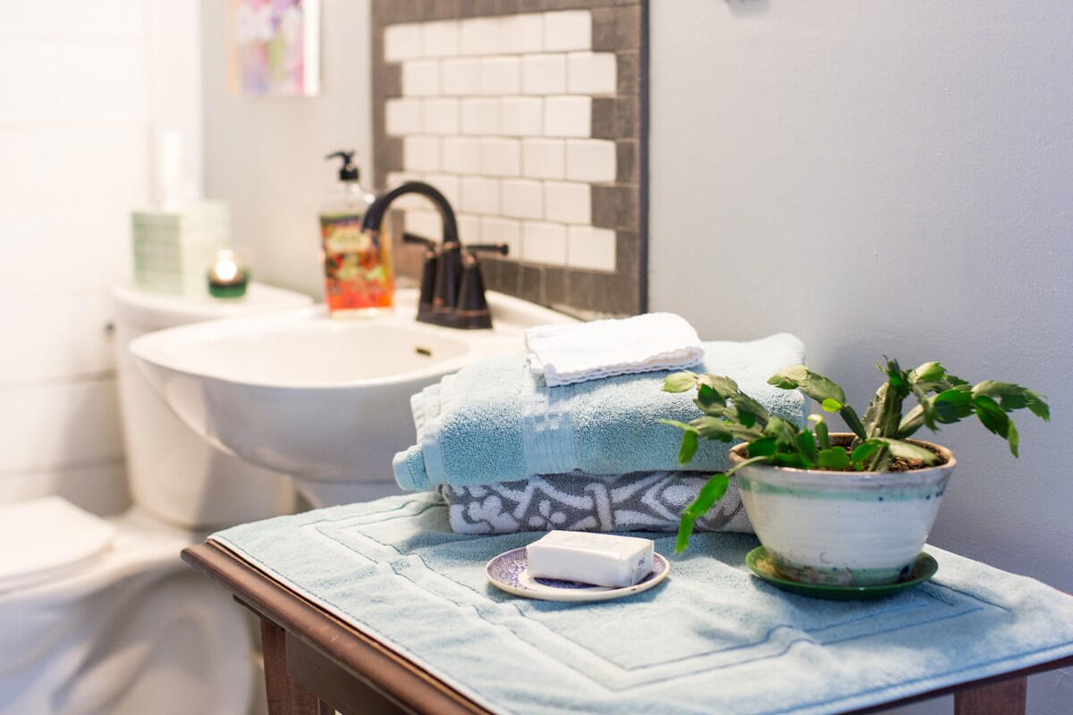 A neat full bathroom displays a white sink and a toilet. Soft, blue towels are neatly stacked beside a small potted plant on a wooden table. A bar of soap and a decorative plate are positioned on the table, adding a touch of charm to the space.
