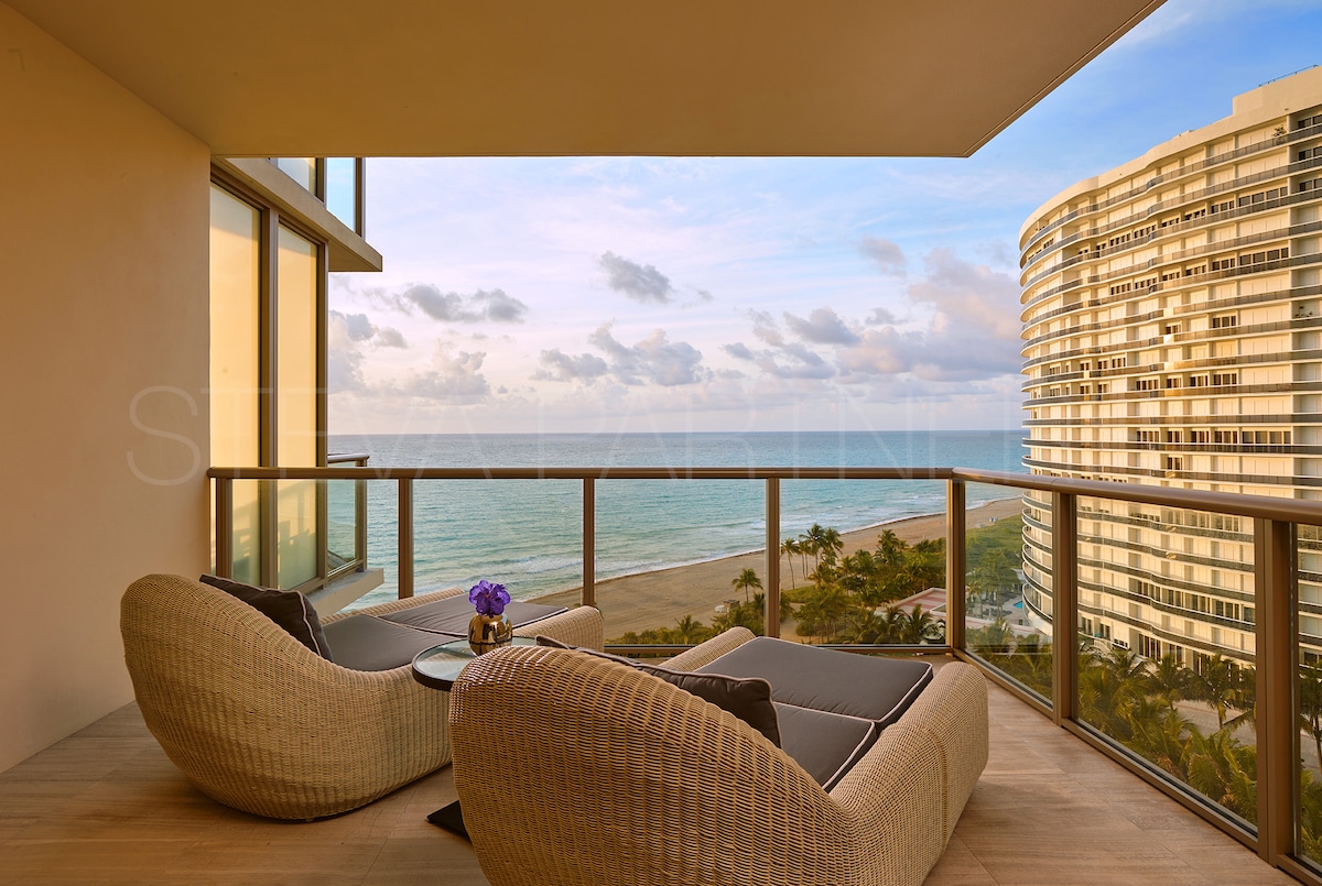 A spacious balcony offers two woven lounge chairs, positioned for ocean views. A small table holds a floral arrangement, while the calm Atlantic stretches beyond. The curved building structure rises in the background, reflecting the soft hues of the sky at dusk.