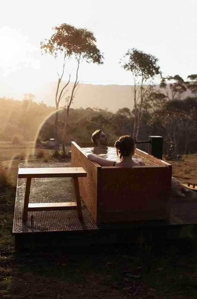 A wooden bath sits on a raised platform, overlooking a serene landscape at sunset. Soft sunlight bathes the scene, casting a warm glow over the trees and rolling hills in the distance, providing an inviting atmosphere for relaxation.