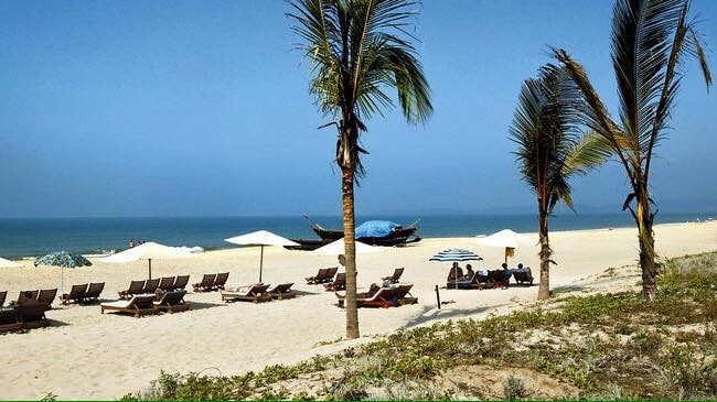 A serene beach scene is depicted, featuring a stretch of soft white sand lined with palm trees. Beach loungers are arranged under umbrellas, while traditional fishing boats are visible on the calm water. The clear blue sky enhances the peaceful atmosphere.