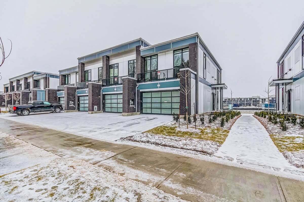 A contemporary marina cottage is shown, featuring a corner unit with a landscaped pathway leading to the entrance. The exterior showcases a mix of dark brick and light panels, with large windows allowing natural light in. A private driveway accommodates vehicles.