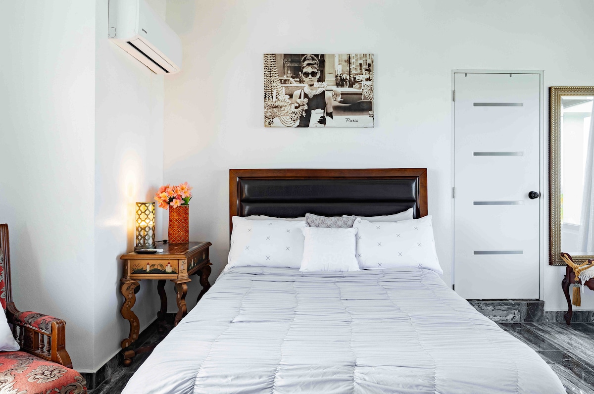 A cozy bedroom features a spacious bed with a light grey duvet and decorative pillows. A vintage side table holds a lamp and a bouquet of faux flowers. A large mirror and a contrasting wooden chair accent the room, while an air conditioning unit is mounted on the wall.