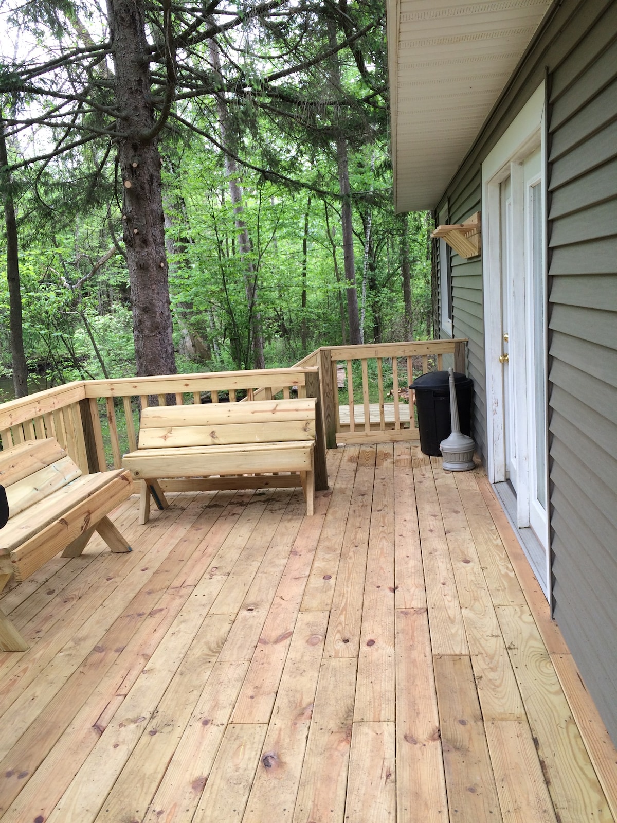 An outdoor deck is seen, featuring wooden seating and a table. The area is surrounded by lush greenery, creating a natural setting. Garbage and a light fixture are positioned on the deck, providing convenience and illumination.