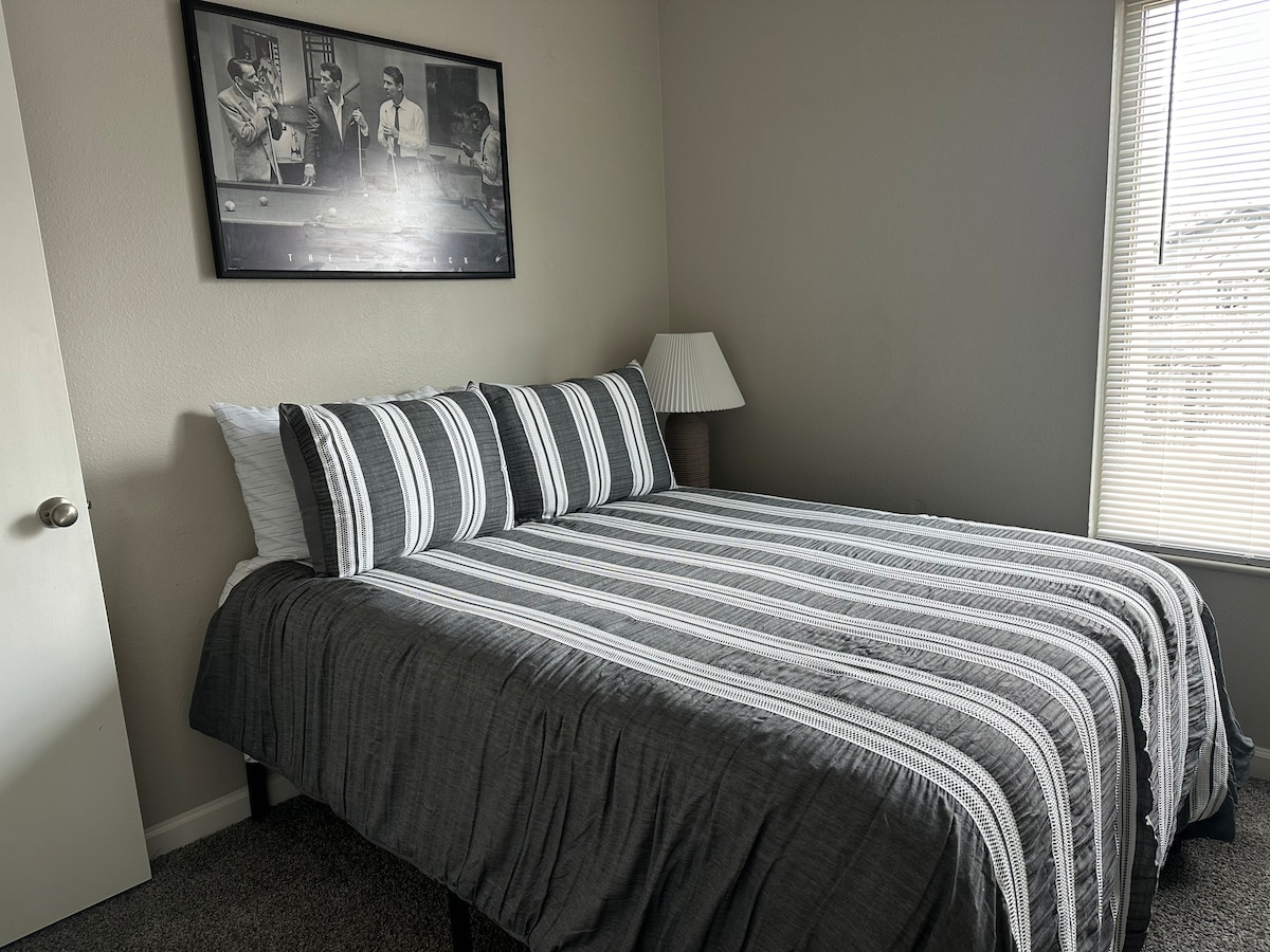 A cozy sleeping area features a full-size bed with a striped gray and white bedspread, accented by patterned pillows. A bedside lamp stands beside the bed, and a large window allows natural light to fill the room, complemented by a minimalist wall art piece.