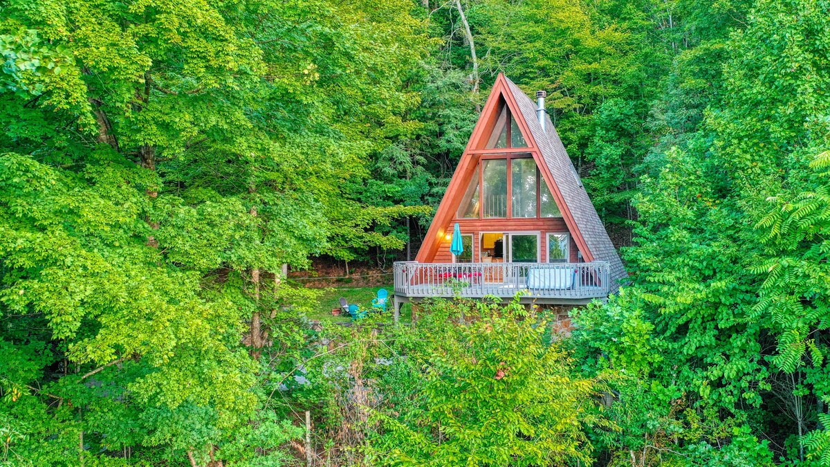 An A-frame cabin is nestled among lush greenery, featuring a triangular roof and large windows. A spacious deck extends from the structure, furnished with chairs. Vibrant trees surround the cabin, creating a serene backdrop of nature.