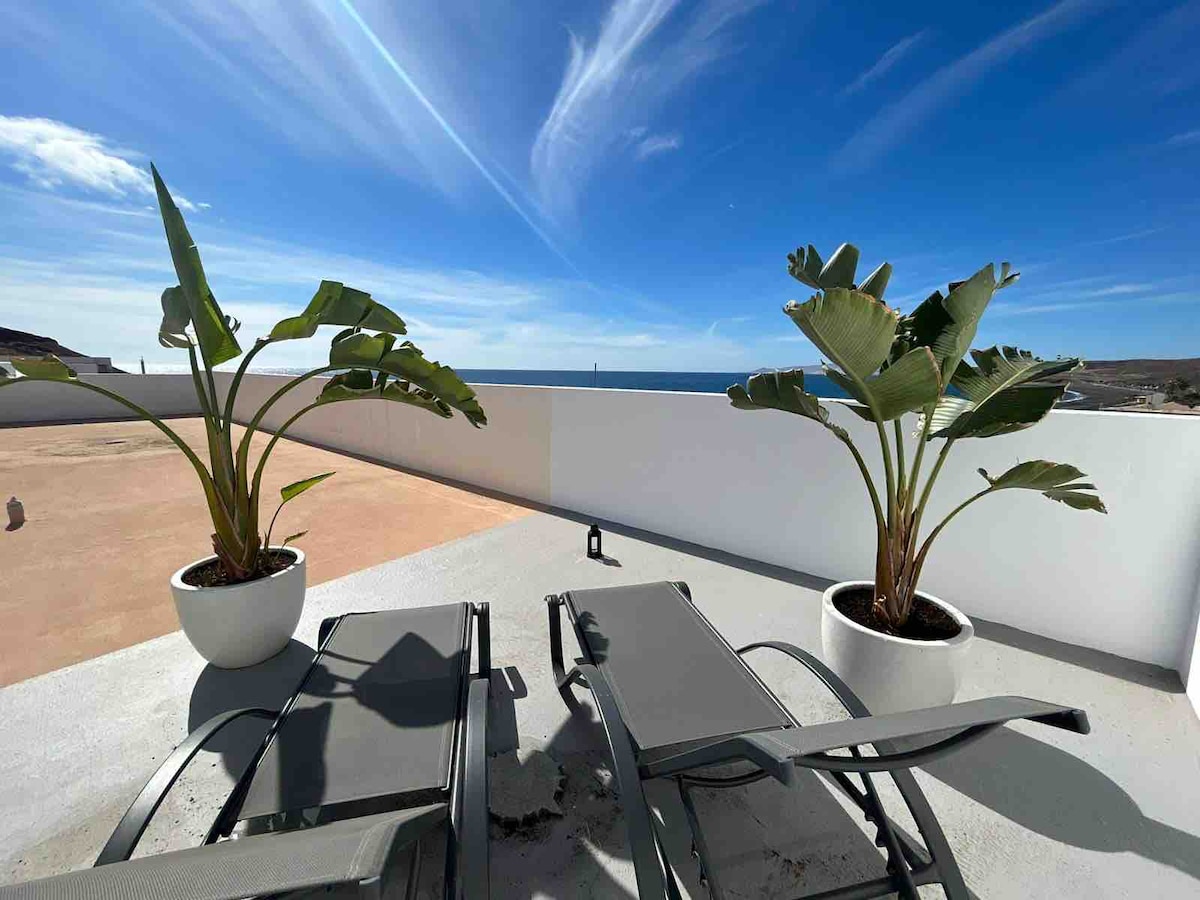 A private solarium is presented with two lounge chairs positioned for relaxation. Potted tropical plants add a natural touch, while clear blue skies and distant sea views create a serene setting. Sunlight brightens the space, enhancing the inviting ambiance.