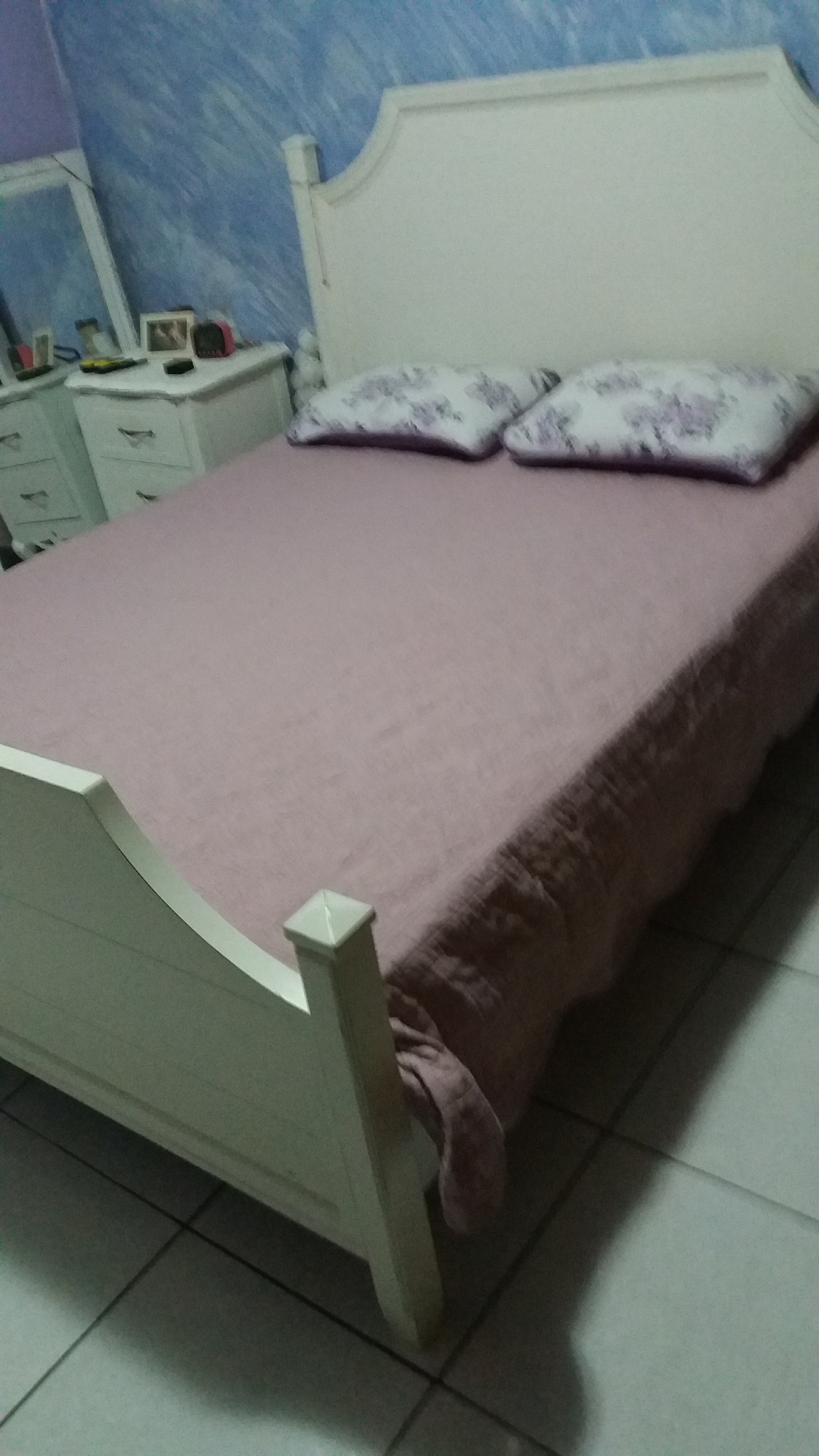 A spacious bed with a light purple bedspread and floral pillows is positioned against a blue wall. A bedside table with small decor items is visible beside the bed. The room features tile flooring and a cozy, restful ambiance.