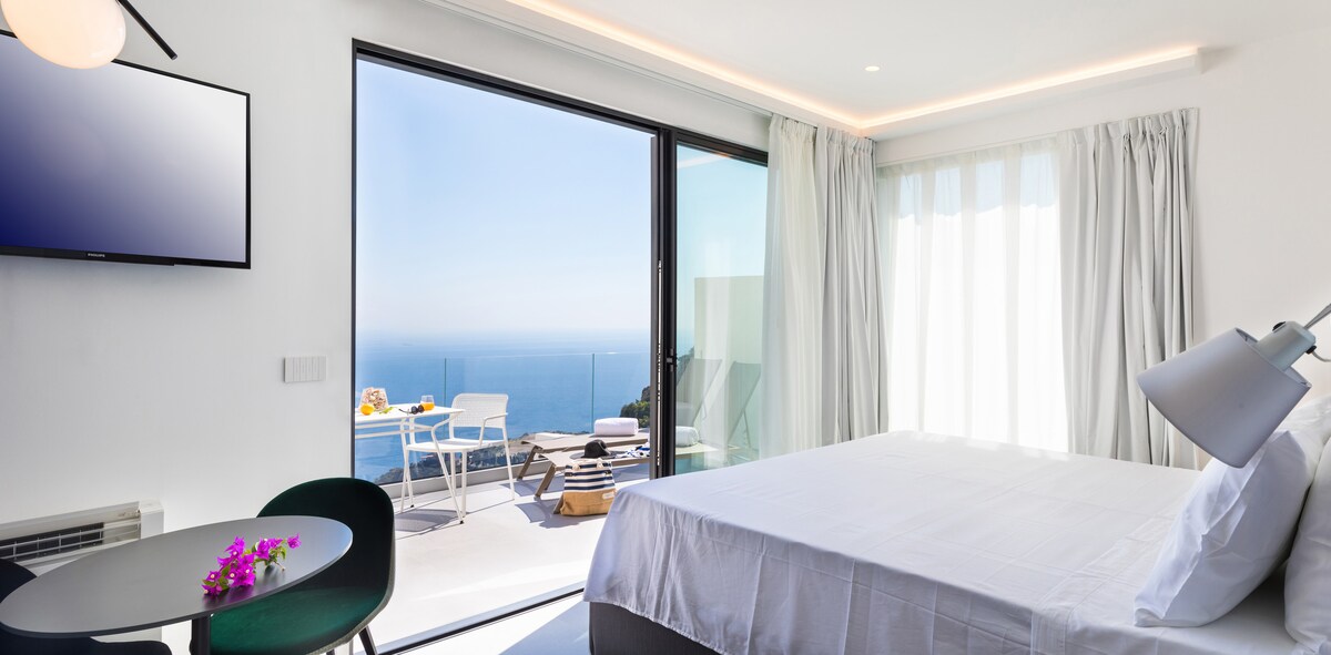 A bright and airy room features a spacious bed with crisp white linens. Large glass doors open to a furnished balcony overlooking the serene sea. A small dining table is set beside a window, with natural light enhancing the minimalist decor.