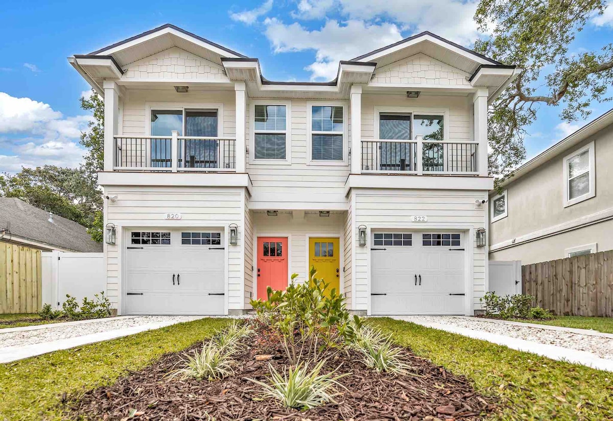 A two-unit beach house is presented in a modern style, featuring a light exterior with contrasting colorful doors. The landscaped front area includes green plantings and a gravel pathway, leading to separate entries for each unit. A serene atmosphere is created by the surrounding trees.