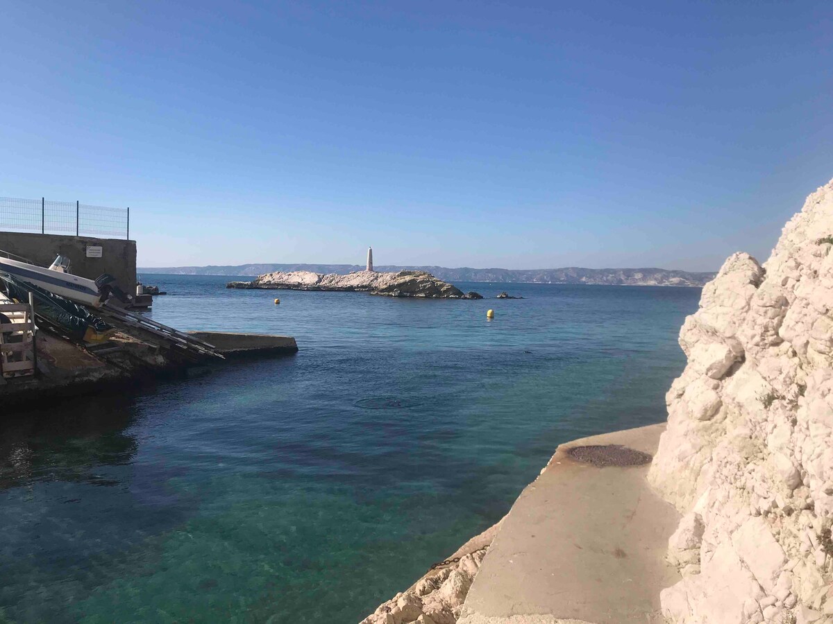 A clear view of calm waters is captured, with rocky shores lining the foreground. A small boat is visible at a wooden dock, and a lighthouse stands on the distant island, surrounded by blue sea and a cloudless sky.