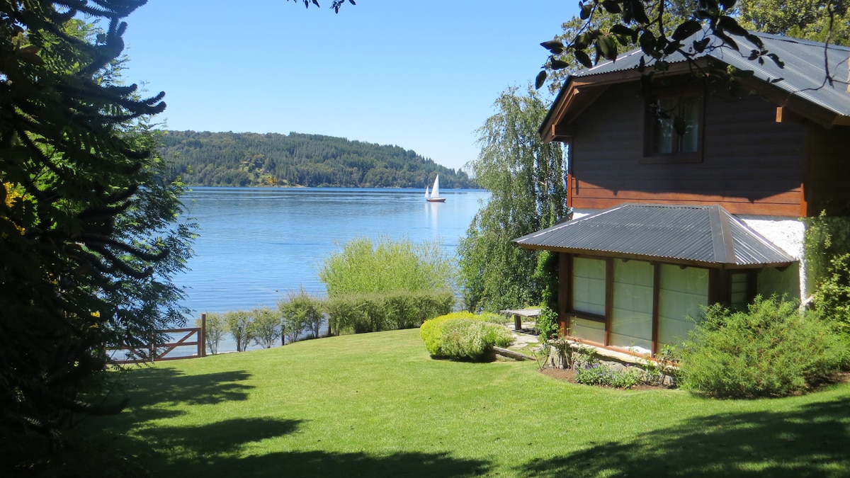 A serene view of the cabin beside a well-maintained lawn leads to a tranquil lakeshore. In the distance, a sailboat glides across the calm waters, framed by lush greenery and tree line. The cabin features large windows that invite natural light.