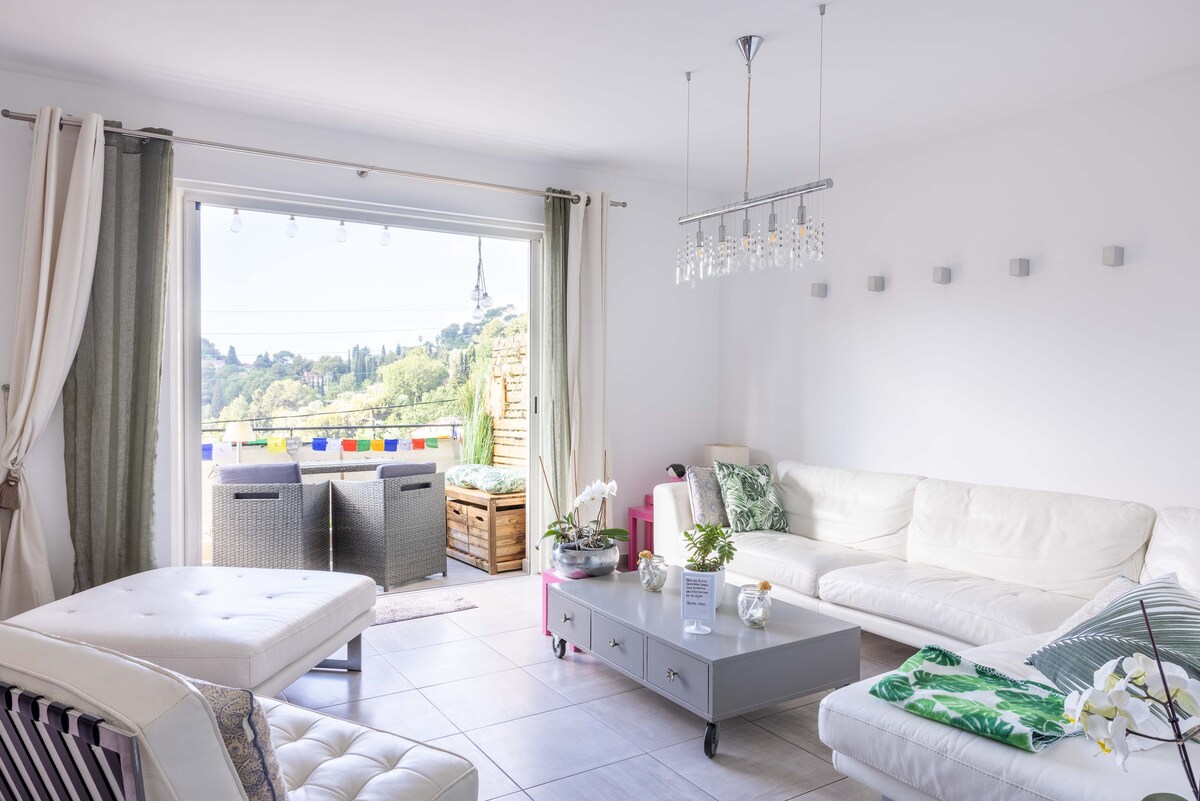 A bright living area features a spacious white sectional sofa and a sleek coffee table. Natural light floods through large glass doors, leading to a balcony with scenic views. Contemporary decor includes a chandelier, potted orchids, and subtle accents of greenery for a calming atmosphere.