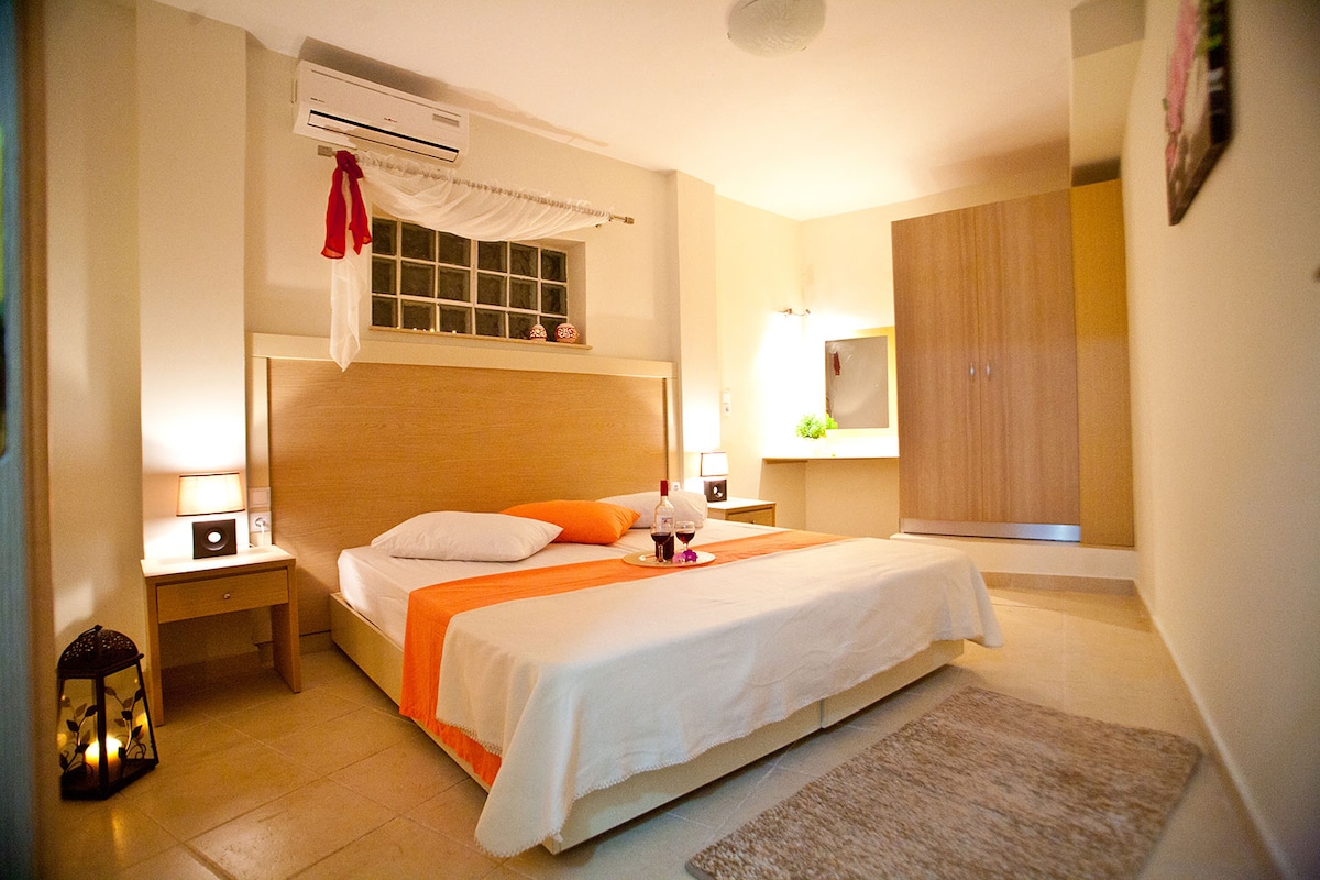 A spacious bedroom features a large bed with a light-colored bedding set and an orange accent. Nightstands with lamps are positioned on either side of the bed. A large wooden wardrobe and a stylish rug complement the simple decor, while a window allows natural light to fill the room.