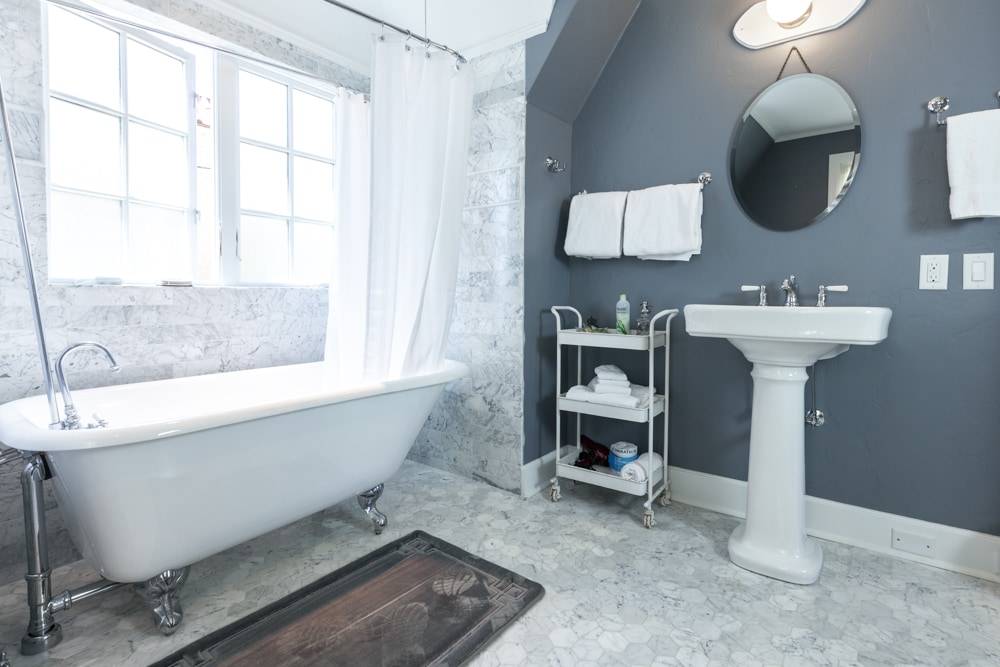 A grand bathroom features a freestanding tub beside a window, allowing natural light to enhance the space. A vintage-style pedestal sink is complemented by a round mirror. Fresh towels are neatly arranged on a wall-mounted rack, with additional linens stored on a rolling cart.