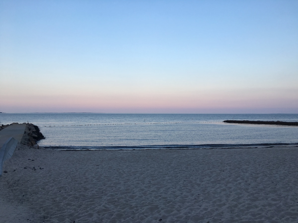 The sandy beach meets calm waters under a pastel sky at dusk. Soft hues of pink and blue are observed across the horizon, and a rocky jetty extends into the water. Gentle waves lap against the shore, creating a serene coastal atmosphere.