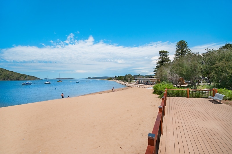 A sandy beach extends along the shoreline, meeting calm blue waters dotted with sailboats. Lush greenery lines the waterfront, with a wooden deck providing a space for relaxation. The sky showcases a mix of white clouds against a vibrant blue backdrop.