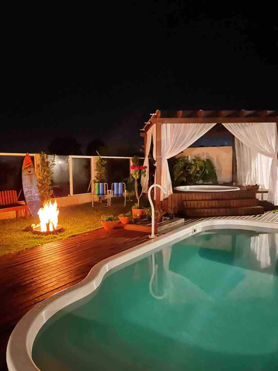 A tranquil outdoor area features a softly lit swimming pool alongside a wooden deck. A jacuzzi is situated under a shaded gazebo, with sheer curtains gently draping. A fire pit is visible, surrounded by potted plants, providing a cozy setting for evening relaxation.