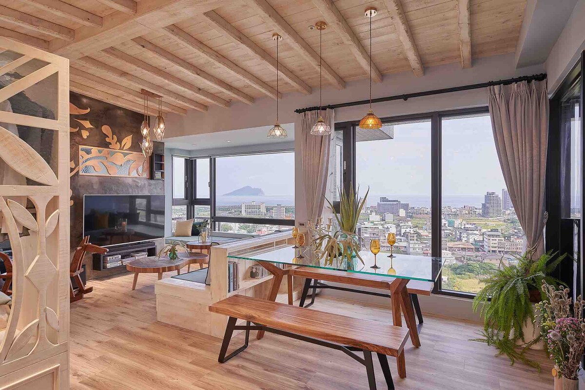 A spacious living area is designed with large windows that offer a view of the ocean and city. The wooden ceiling complements the natural light, while a glass dining table is paired with wooden benches. Green plants are placed near the windows, enhancing the space.