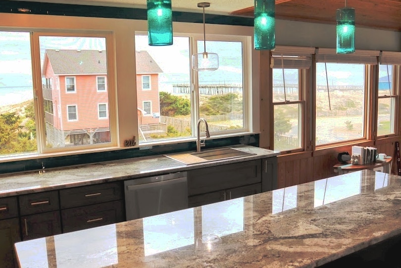 A well-equipped kitchen features a spacious granite countertop and views of the ocean through large windows. The area is illuminated by unique teal pendant lights, and the sink is positioned to take advantage of the scenic backdrop. The serene setting allows for natural light to enhance the space.