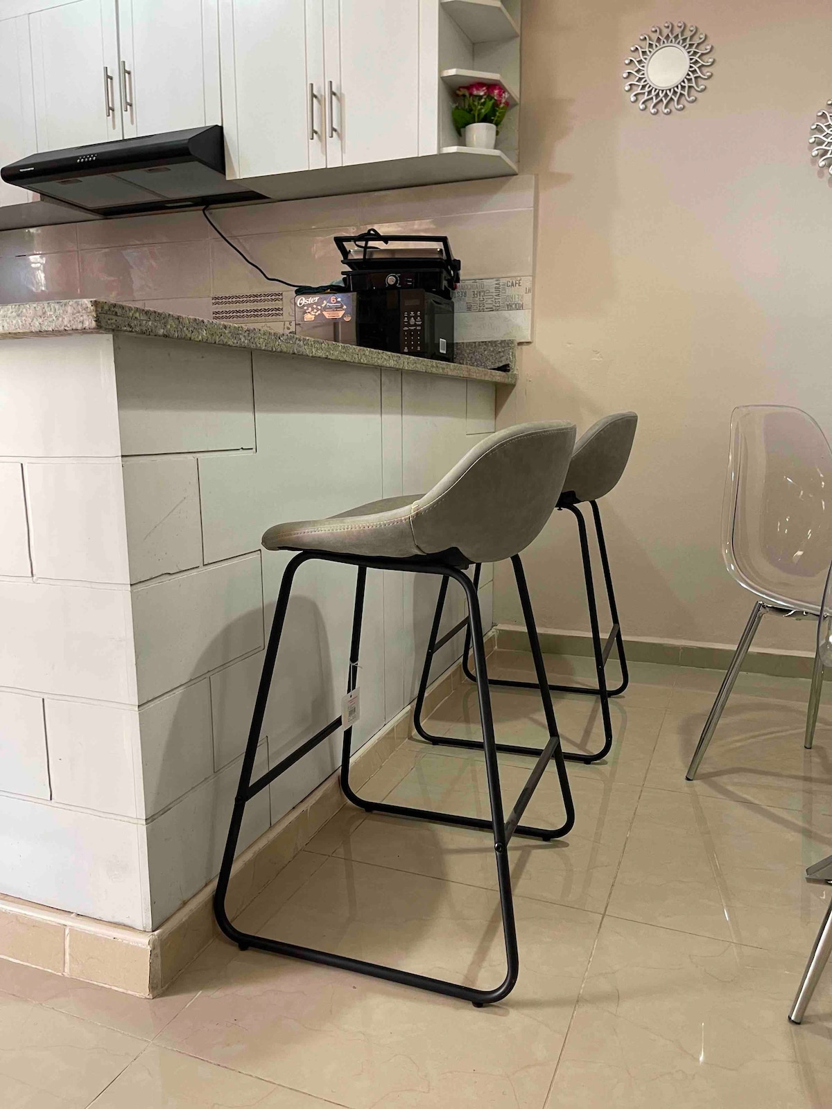 Two modern bar stools with soft, cushioned seats are positioned at a kitchen counter made of light-colored granite. A coffee maker and some kitchen supplies are visible on the countertop, complemented by a small plant and decorative mirrors on the wall.