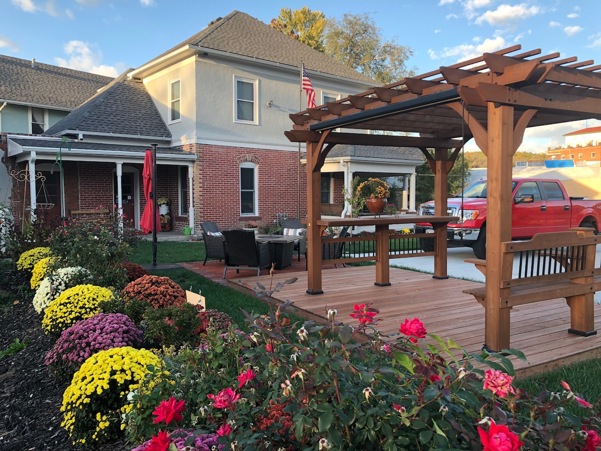 A cozy outdoor space features a wooden pergola with seating, surrounded by vibrant flower beds filled with various colorful blooms. The inviting brick facade of the building is visible in the background, and a flag is displayed, adding to the welcoming ambiance.