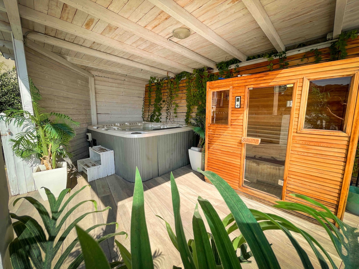 A private relaxation area features a spacious hot tub surrounded by lush greenery. A sauna with wooden panels is visible to the right, offering a cozy ambiance. The area is well-lit, showcasing a modern design with wood accents and potted plants enhancing the tranquil environment.